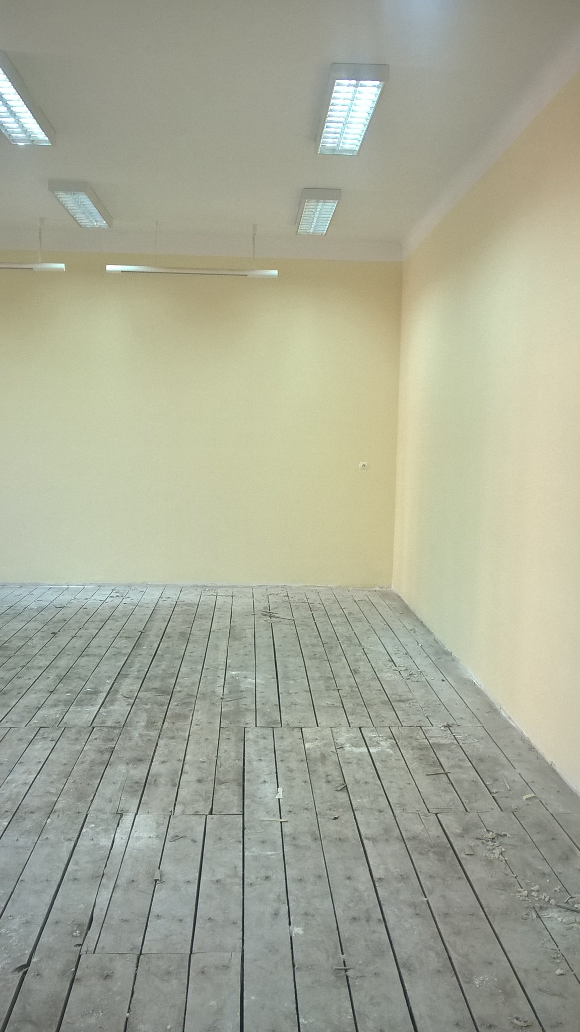Pusta, jasna przestrzeń z pomalowanymi na kremowo ścianami i starym, drewnianym parkietem wymagającym renowacji, oświetlona przez podłużne lampy sufitowe.