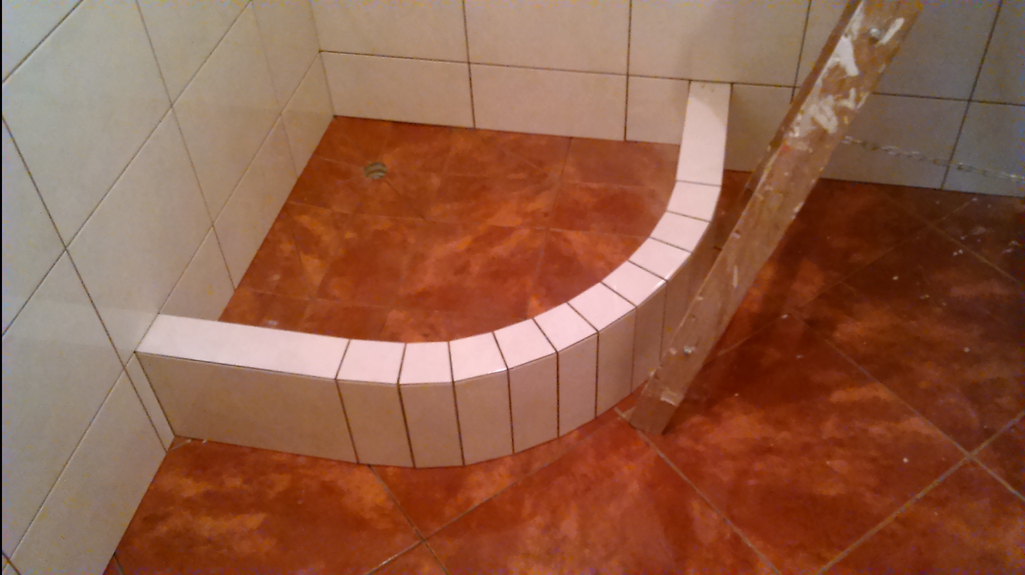 Niedokończona kabina prysznicowa z brązową podłogą i białą, półokrągłą obudową z płytek, widoczne ślady zaprawy i drabina oparta o ścianę.