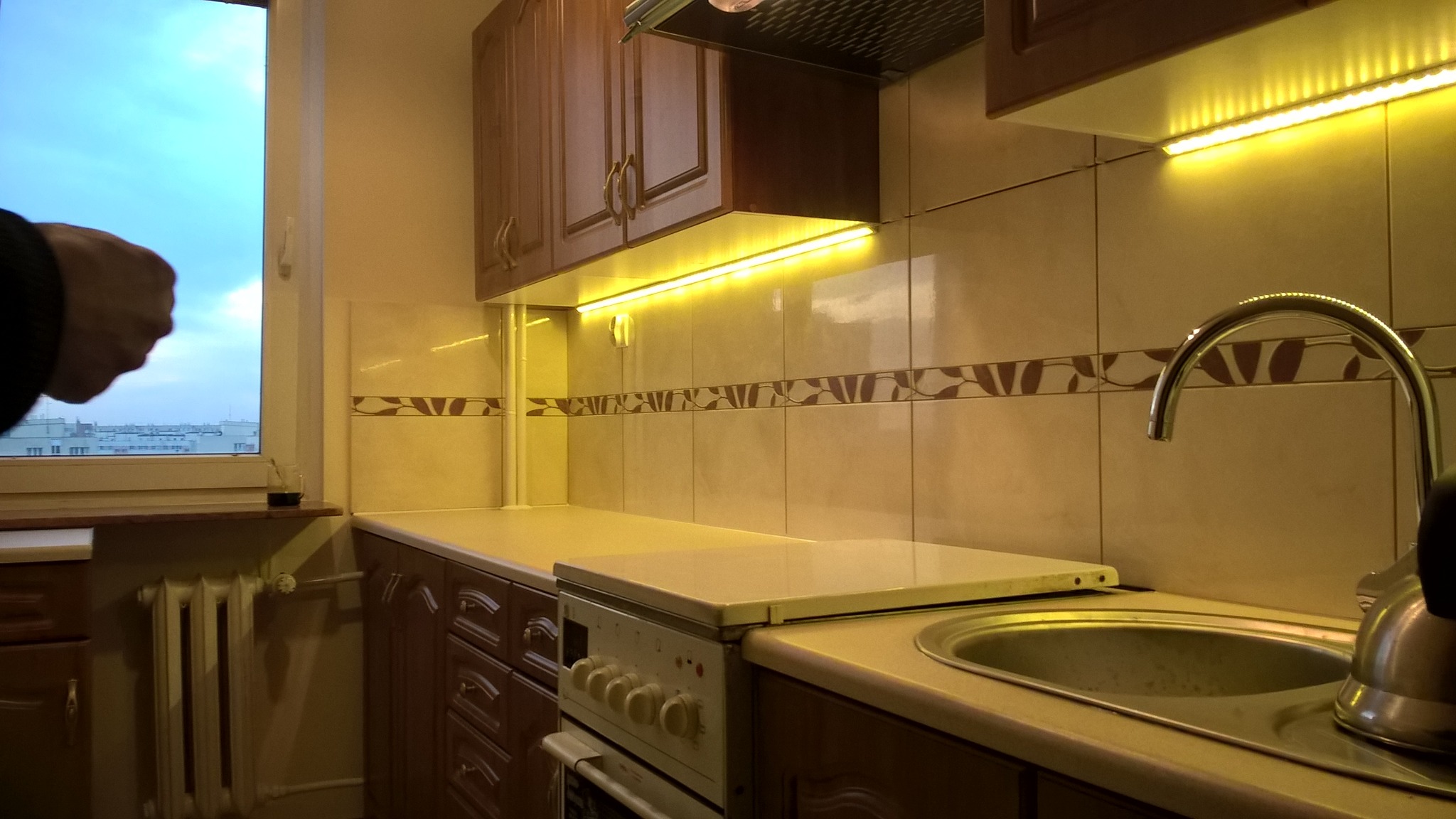 Wykończona kuchnia z brązowymi szafkami, beżowymi płytkami z ciemnobrązowym wzorem i oświetleniem LED pod szafkami, z widocznym oknem i fragmentem dłoni w lewym górnym rogu.