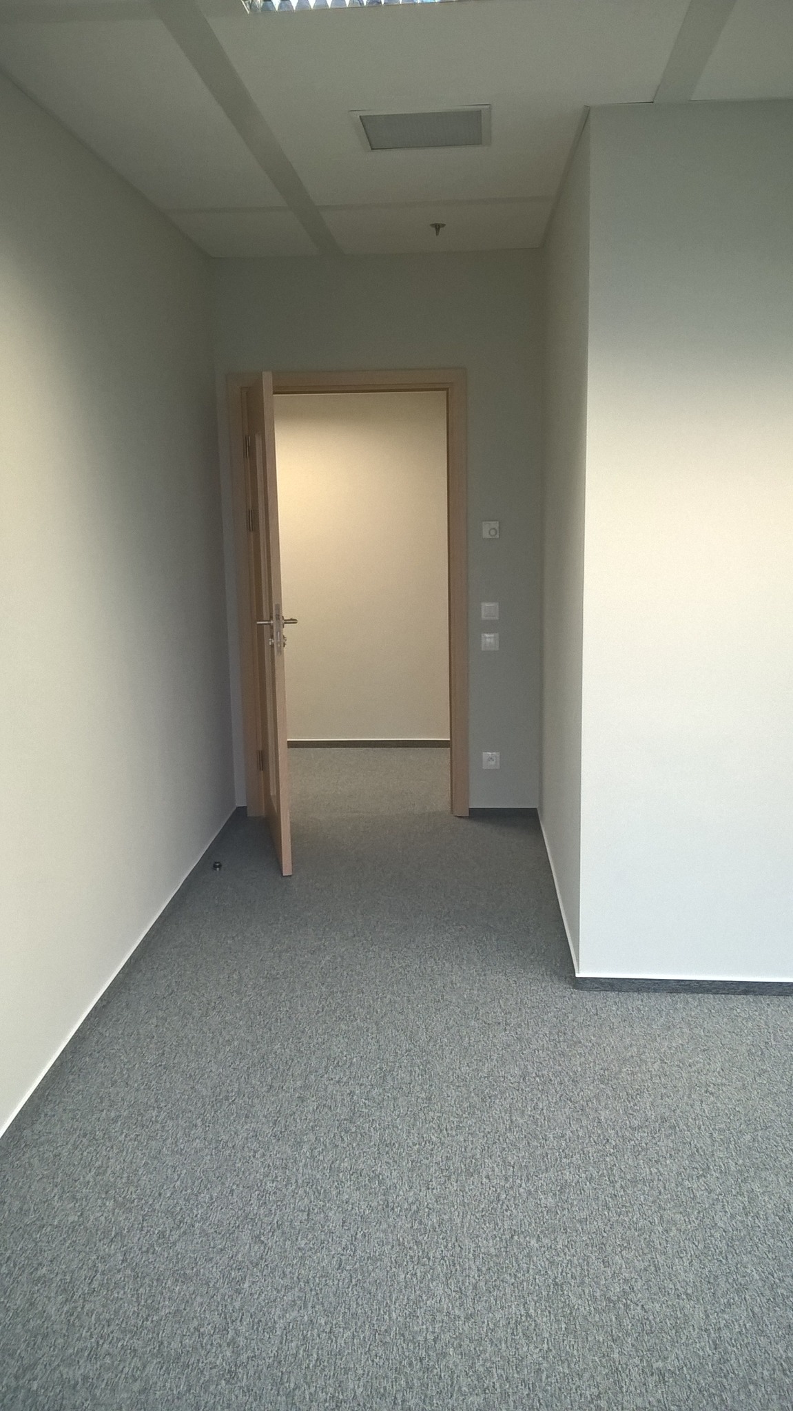 Pomalowane na szaro ściany biura z szarym dywanem, otwarte drzwi do sąsiedniego pomieszczenia, widoczne włączniki światła.