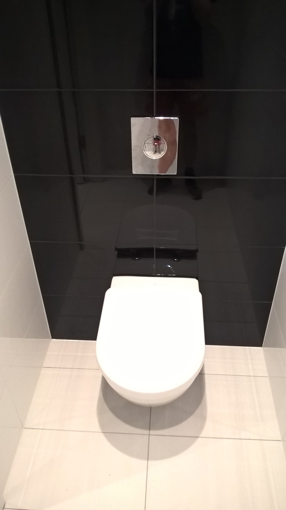 Nowoczesna toaleta podwieszana z białym siedziskiem i chromowanym przyciskiem spłukiwania na tle czarnych płytek, uzupełnona jasnymi płytkami podłogowymi, widok z góry.