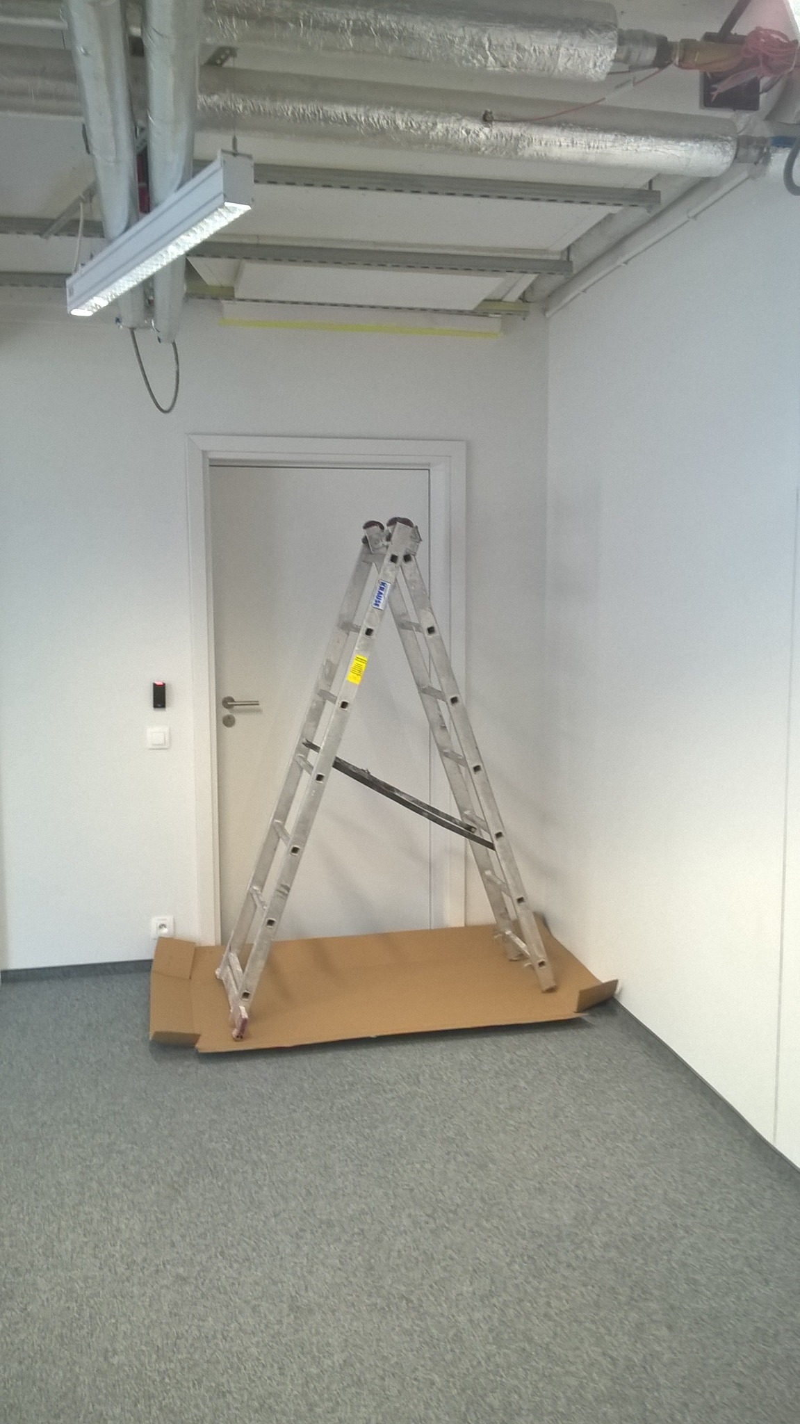 Wnętrze biura przed remontem: drabina oparta o ścianę, zabezpieczona kartonem, widoczne instalacje pod sufitem i oświetlenie LED.
