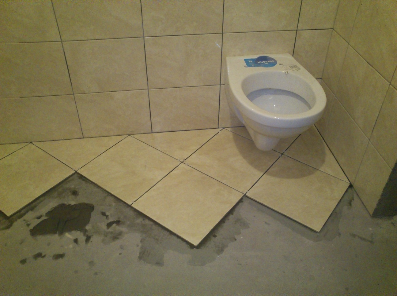 Biała toaleta wisząca na tle częściowo wyłożonej podłogi beżowymi, kwadratowymi płytkami ułożonymi w karo, widoczny surowy beton pod płytkami.