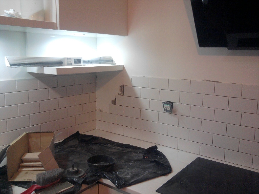 Białe, prostokątne płytki ułożone na ścianie w kuchni, widoczne narzędzia i materiały budowlane, fragment niezabezpieczonego gniazdka elektrycznego.