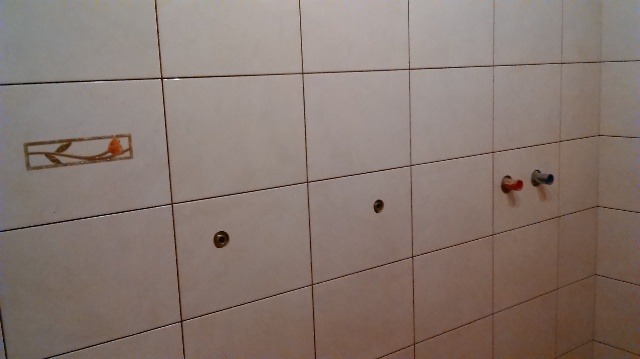 Ściana w łazience wyłożona białymi płytkami z dekoracyjnym motywem kwiatowym i przygotowanymi otworami na armaturę oraz wystającymi rurami zasilającymi ciepłą i zimną wodę.