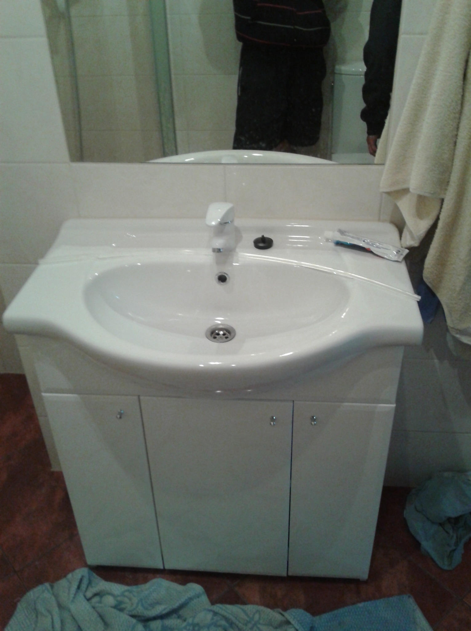 Biała umywalka z szafką w łazience, widoczna tuba pasty do zębów, odbicie osoby w lustrze, ręcznik i ubrania na podłodze.