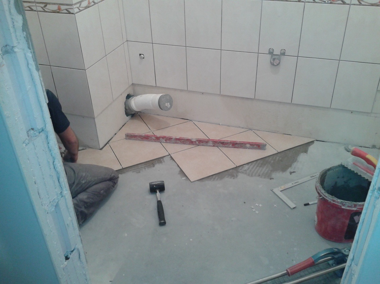 Układanie beżowych płytek podłogowych w łazience, widoczny fragment instalacji kanalizacyjnej, poziomica, młotek i wiadro z zaprawą.