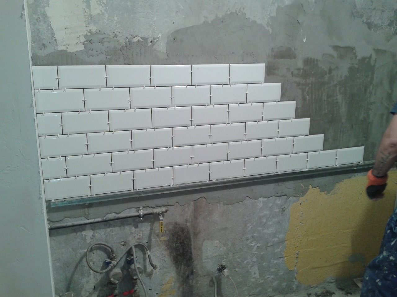 Układanie białych, prostokątnych płytek na ścianie z widocznymi rurami i fragmentami starej farby, użycie krzyżyków dystansowych.