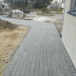 Ułożona, szara kostka brukowa tworzy chodnik. W tle sterta kostki, ziemia i budynek. Widoczny fragment budynku z szarą elewacją.