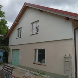 Dwukondygnacyjny dom z ocieploną górną elewacją w jasnym kolorze, kontrastującą z dolną częścią. Widoczna drabina i palety na zewnątrz, sugerujące trwające prace.