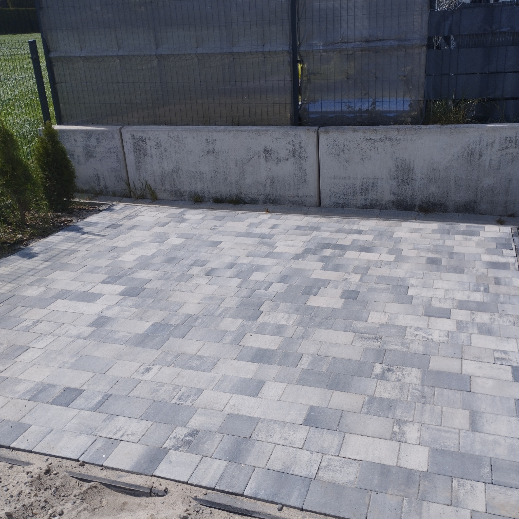 Ułożona kostka brukowa w odcieniach szarości, widok z góry. W tle betonowy murek i siatka ogrodzeniowa. Częściowo widoczna zieleń trawnika.