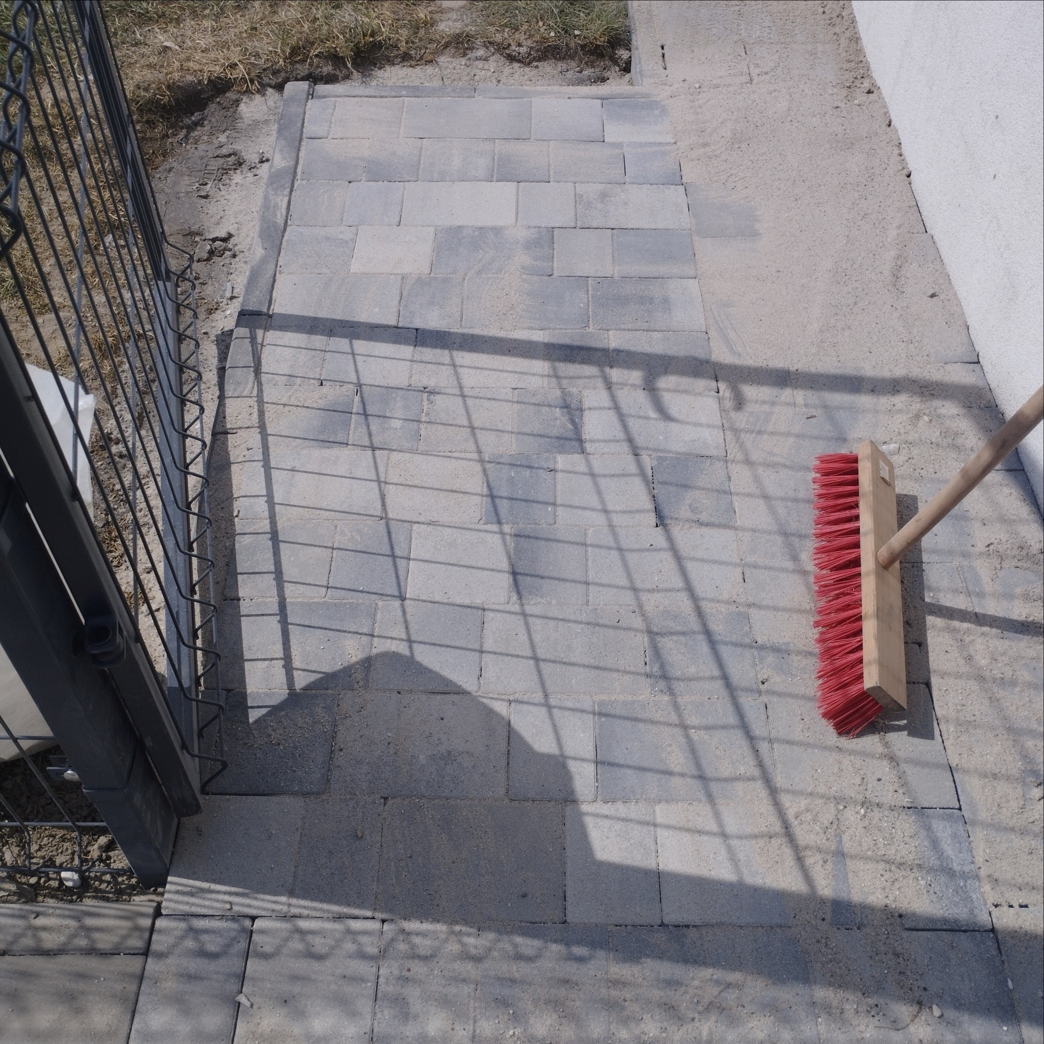 Świeżo ułożona, szara kostka brukowa tworzy ścieżkę. Czerwona zmiotka leży na powierzchni, a cień metalowej furtki pada na bruk. Widoczne drobne ziarna piasku.