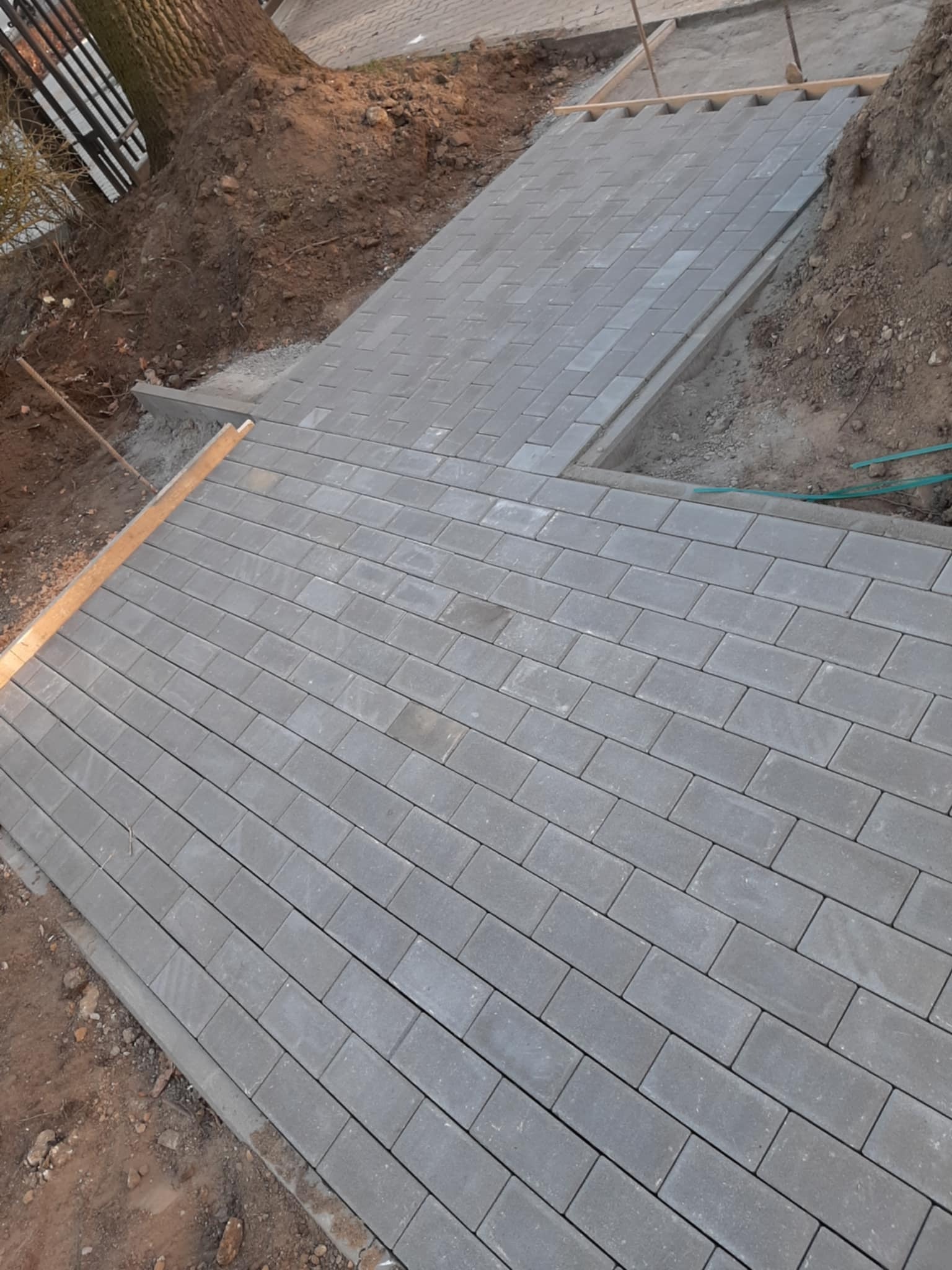 Świeżo ułożona, szara kostka brukowa tworząca ścieżkę ogrodową z obrzeżami. Widoczne przygotowane podłoże i elementy drewniane. Ujęcie z góry, w dziennym świetle.