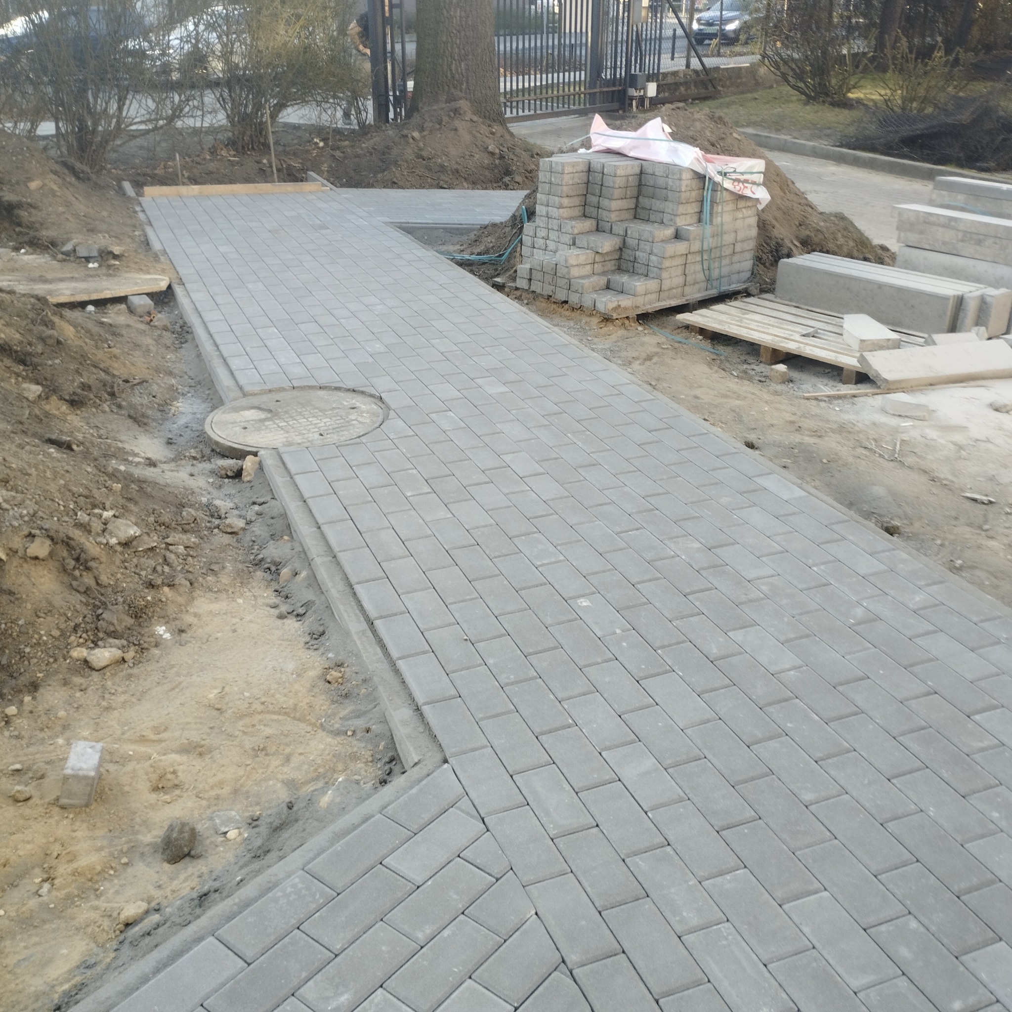 Świeżo ułożona, szara kostka brukowa tworzy chodnik. W tle paleta z kostką i ziemia. Widoczny właz kanalizacyjny wkomponowany w ścieżkę.
