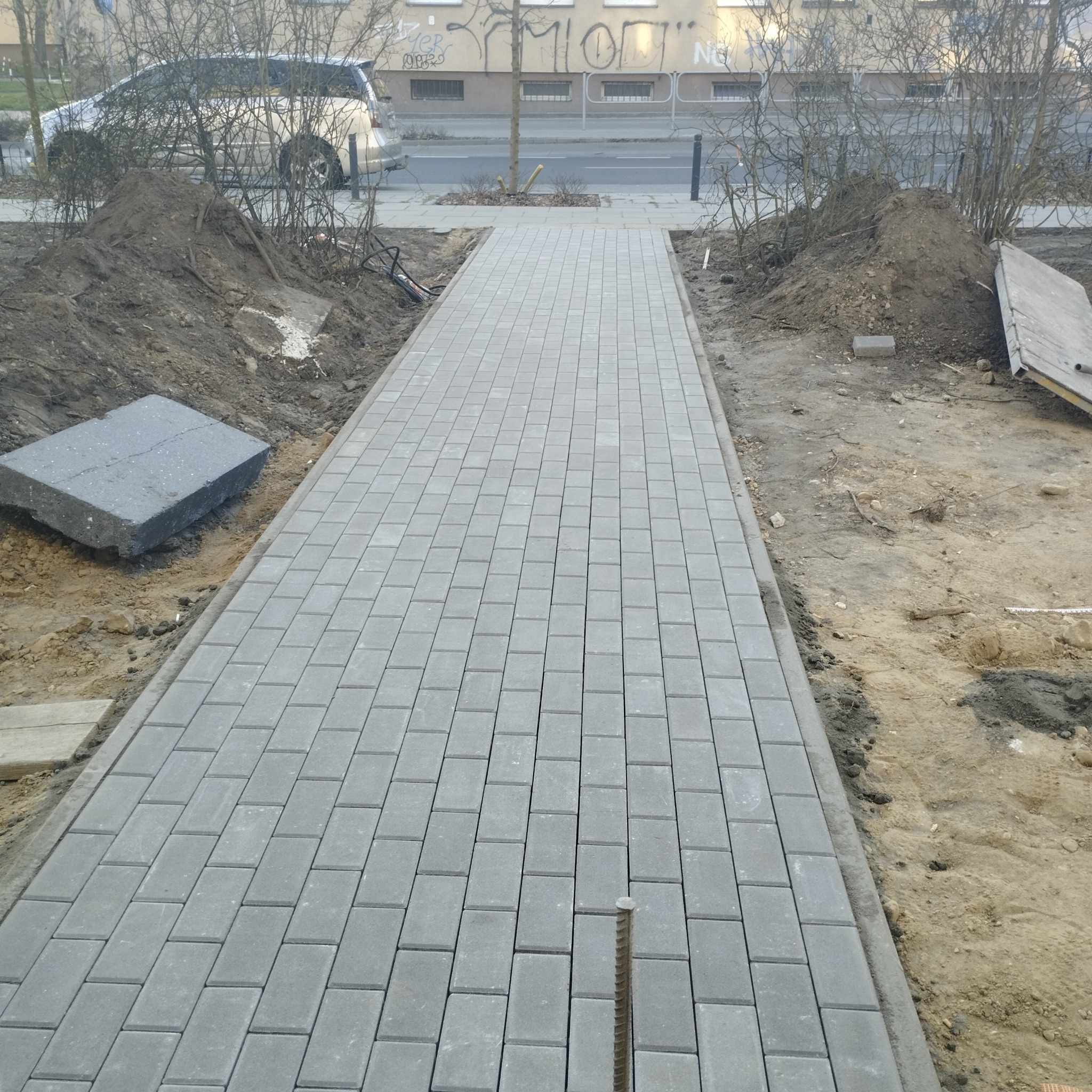 Świeżo ułożona, szara kostka brukowa na ścieżce, z widoczną stertą ziemi i pojedynczymi kostkami obok. Ujęcie z perspektywy pieszego.