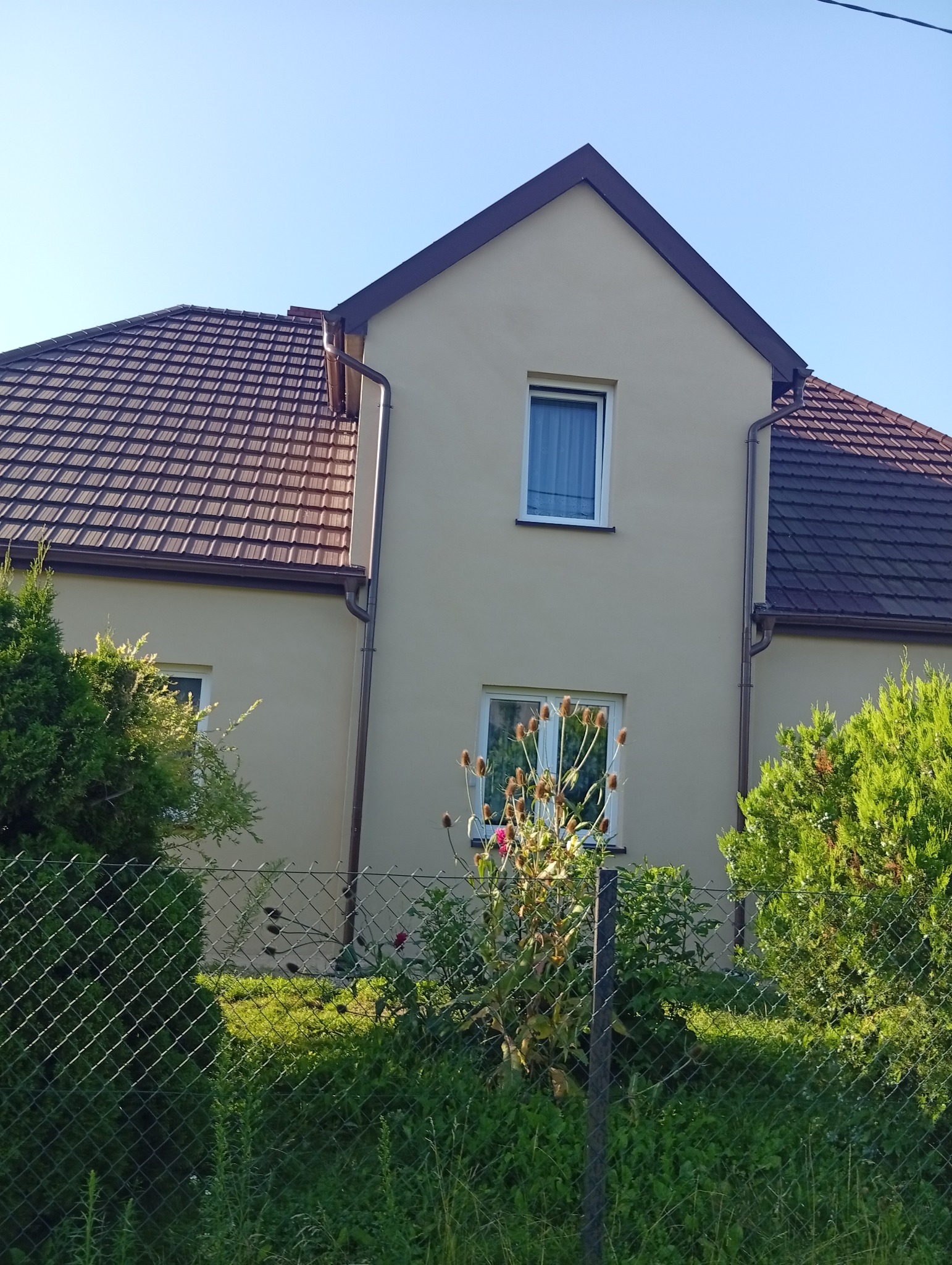 Jasna elewacja dwukondygnacyjnego domu z brązowym dachem, widoczna zza zieleni i metalowego ogrodzenia. Ujęcie w słoneczny dzień.