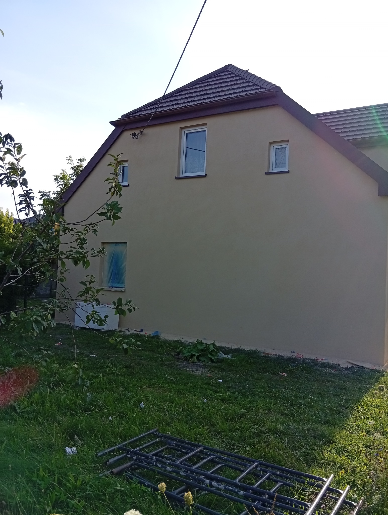 Elewacja domu po remoncie z beżową farbą. Jedno okno zabezpieczone folią. Na trawie leżą metalowe rusztowania. Widok z boku na dom z brązowym dachem.