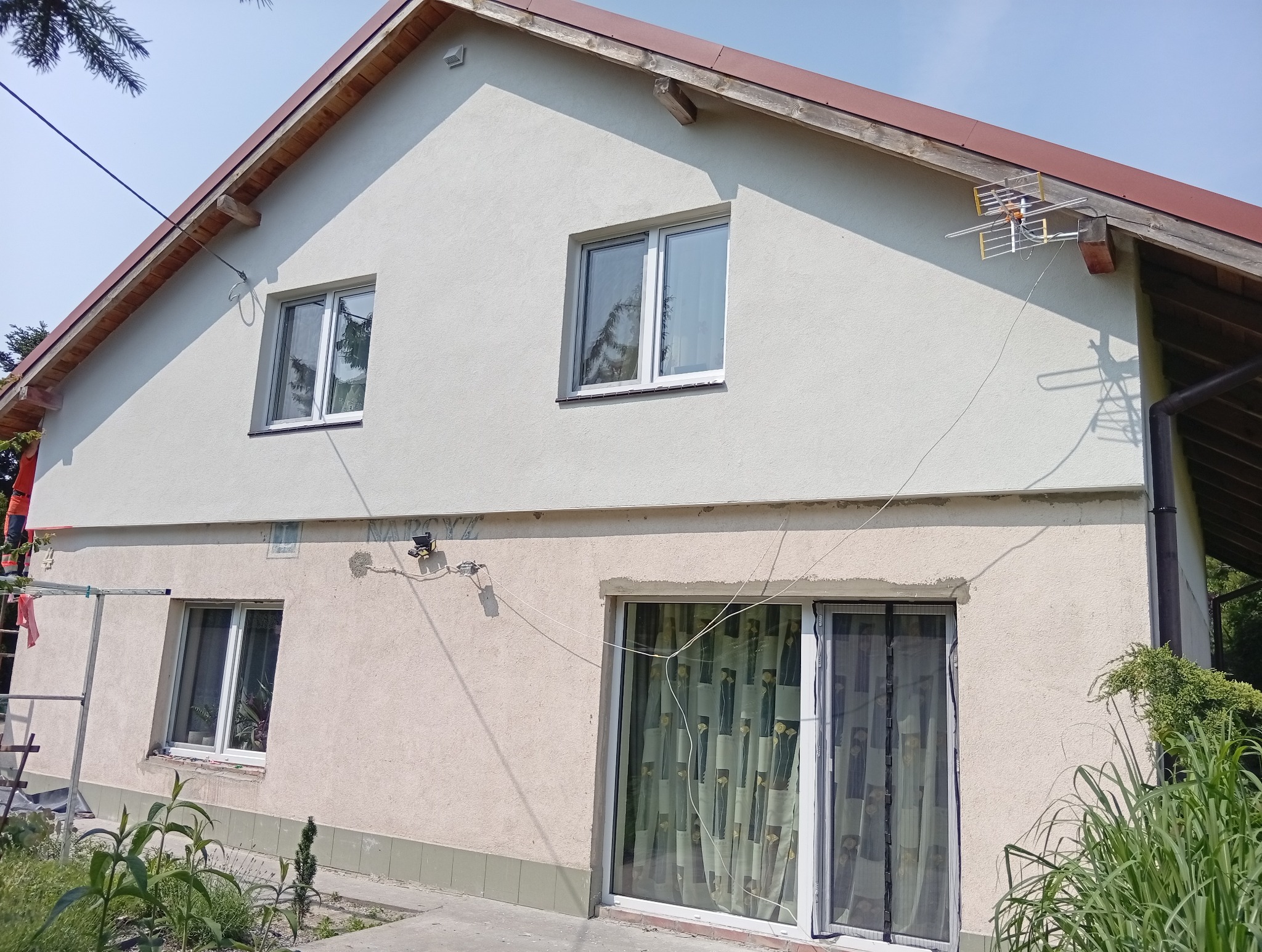 Dwukondygnacyjny dom z ocieploną elewacją w odcieniach beżu, widoczna antena na dachu i fragment rusztowania po lewej stronie. Część elewacji jest jeszcze nieocieplona.