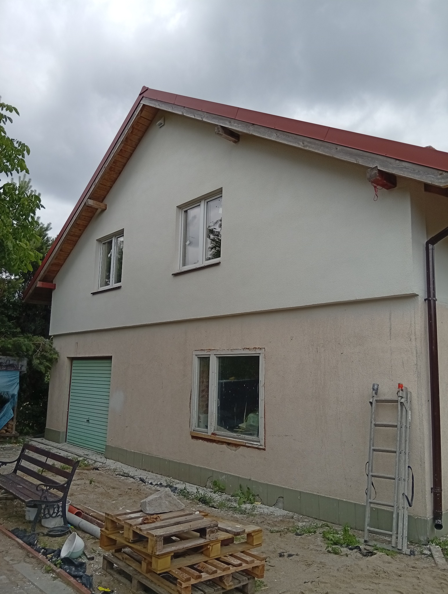 Dwukondygnacyjny dom z ocieploną górną elewacją w jasnym kolorze, kontrastującą z dolną częścią. Widoczna drabina i palety na zewnątrz, sugerujące trwające prace.