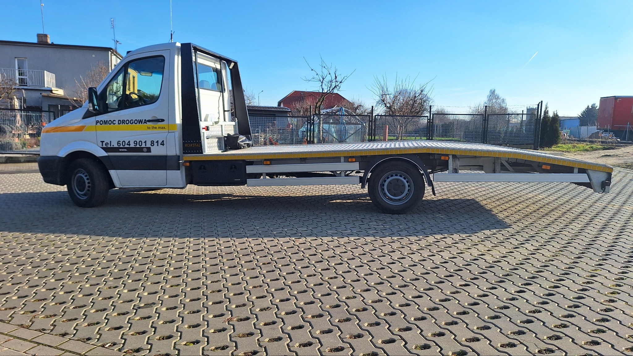 Biała laweta pomocy drogowej z żółtym pasem, zaparkowana na kostce brukowej w słoneczny dzień. Widoczny numer telefonu i napis 'Pomoc Drogowa'.