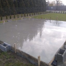 Kompleksovo Sp. z o.o. - Świeżo wylana, gładka płyta betonowa na zewnątrz, z drewnianym szalunkiem i betonowymi bloczkami po bokach, odblaski drzew na powierzchni, w tle trawnik i żywopłot.