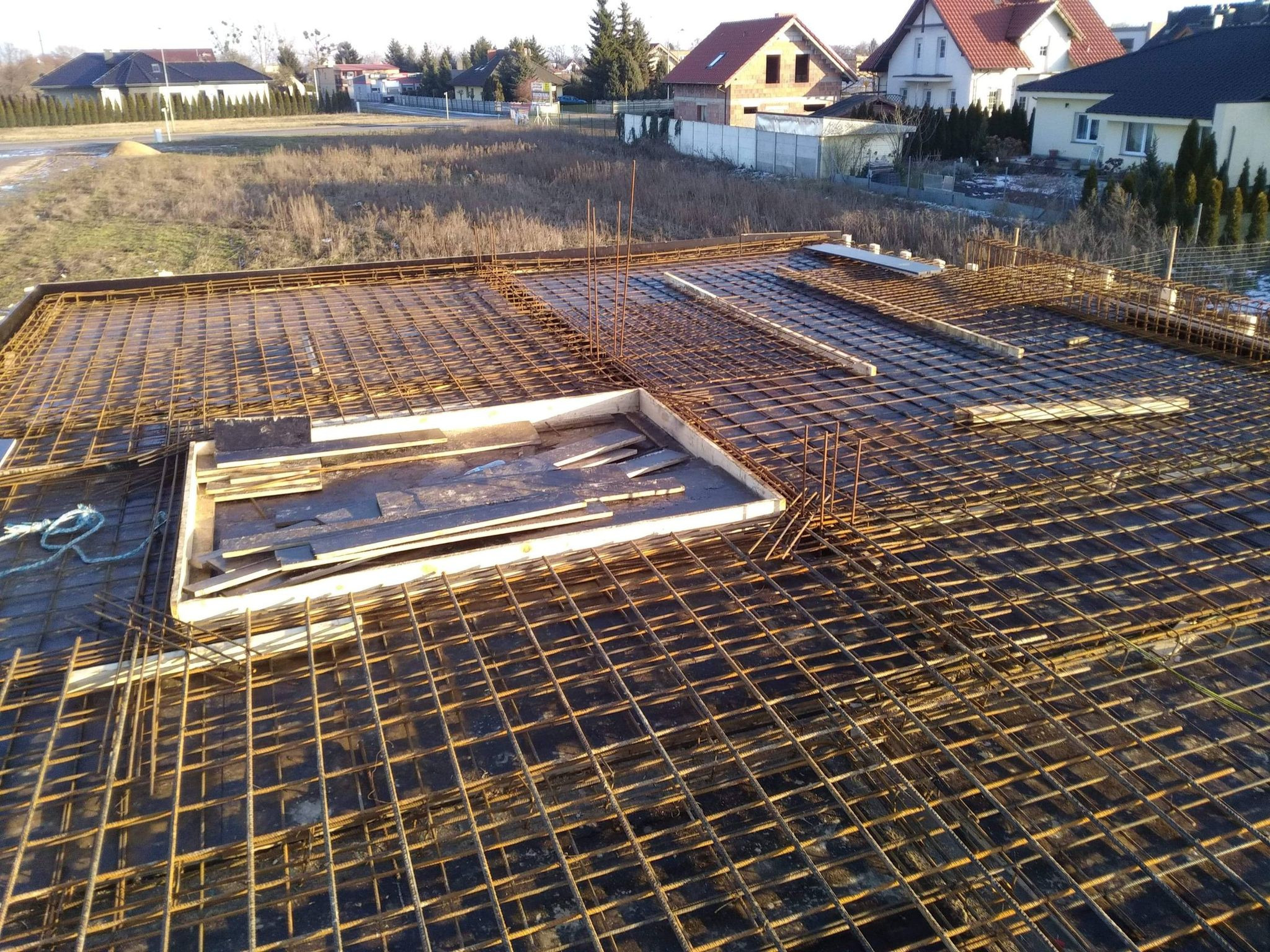 Zbrojenie stropu na budowie domu jednorodzinnego, widok z góry na siatkę z prętów stalowych i szalunki, w tle zabudowa jednorodzinna.