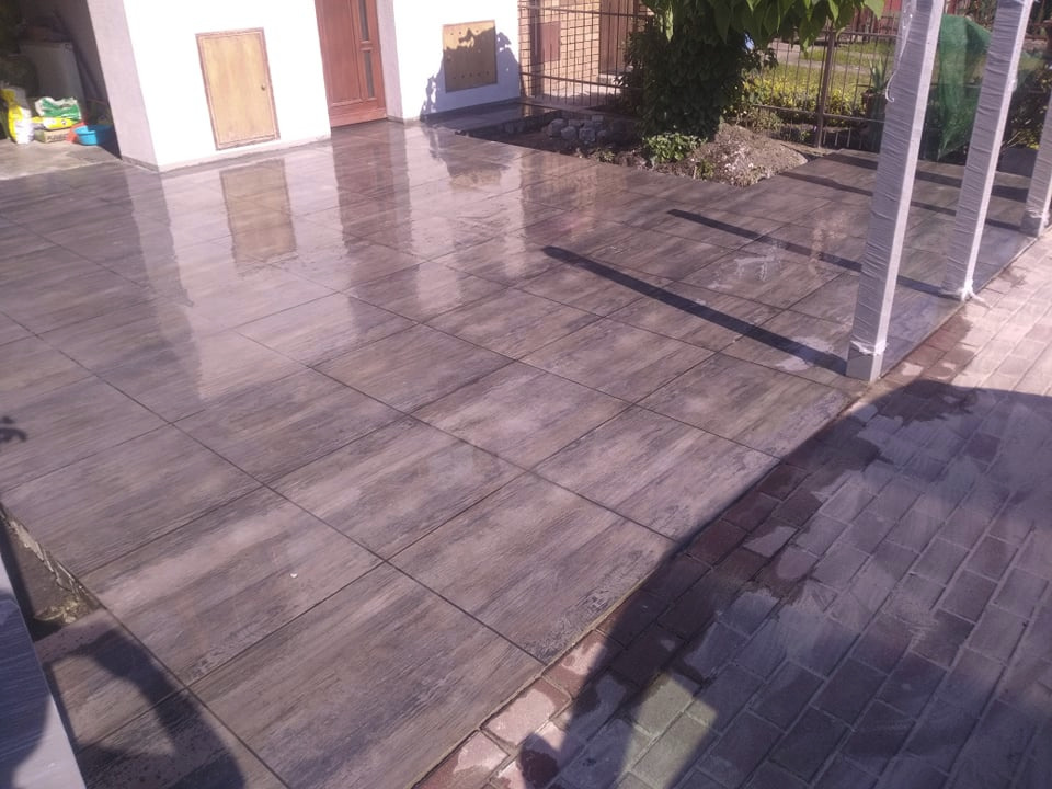 Nowo ułożona, błyszcząca posadzka z płyt imitujących drewno na zewnątrz budynku, widoczne refleksy światła, obrzeże z kostki brukowej.