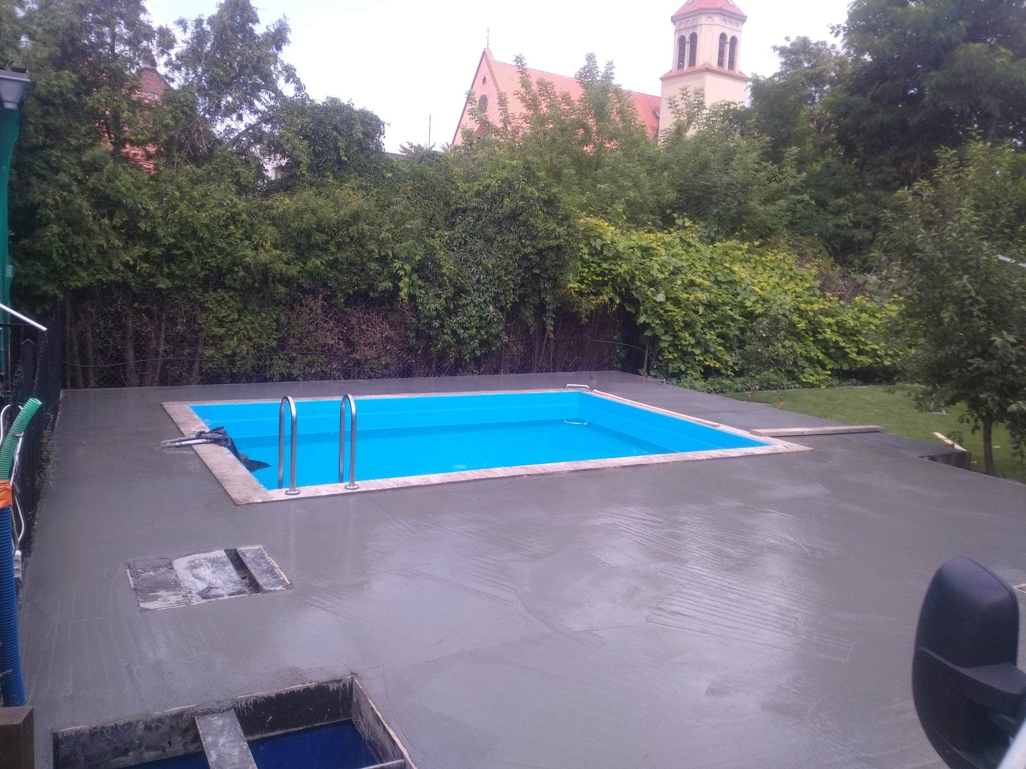 Świeżo wylana, jeszcze mokra, betonowa wylewka wokół basenu z widocznymi włazami rewizyjnymi i metalową drabinką basenową, w tle zieleń i budynek z wieżą.