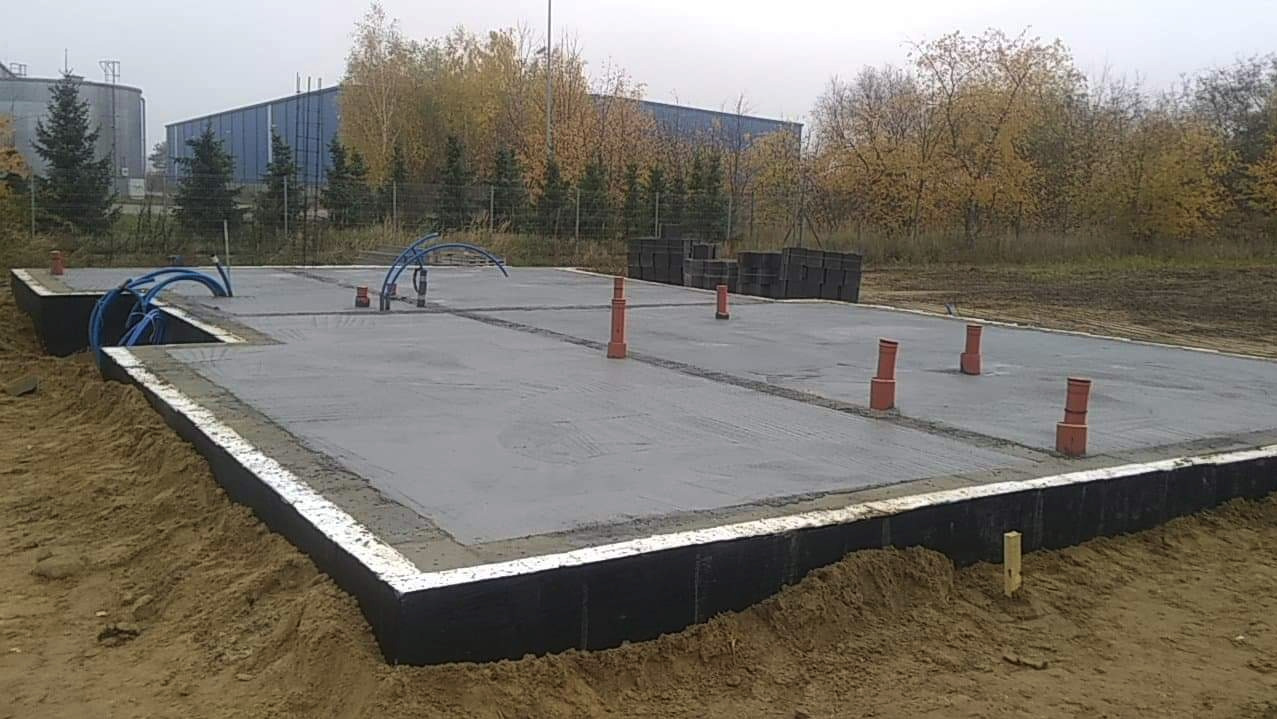 Świeżo wylana, gładka betonowa płyta fundamentowa z wystającymi rurami kanalizacyjnymi i niebieskimi przewodami, zabezpieczona czarną izolacją, otoczona piaskiem, z widocznym budynkiem przemysłowym...