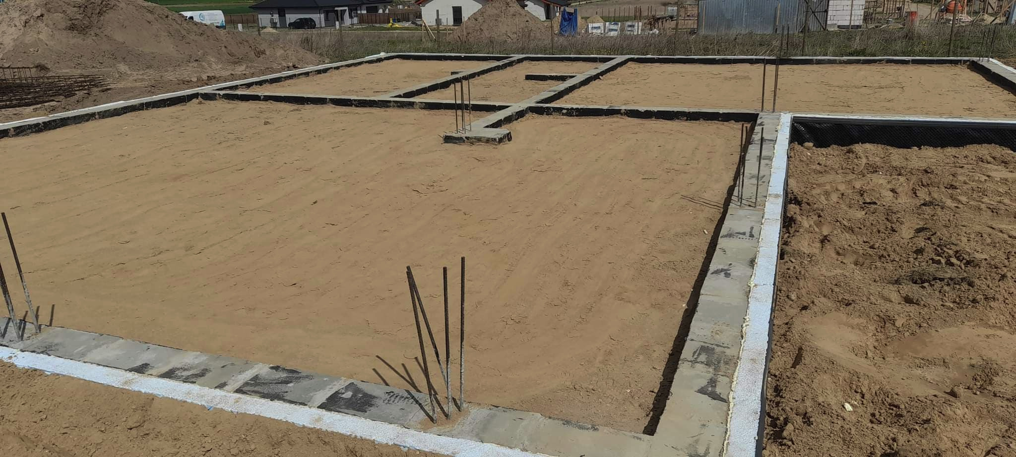 Przygotowanie fundamentów pod budowę domu, z widocznymi zbrojeniami i izolacją, teren wyrównany piaskiem.
