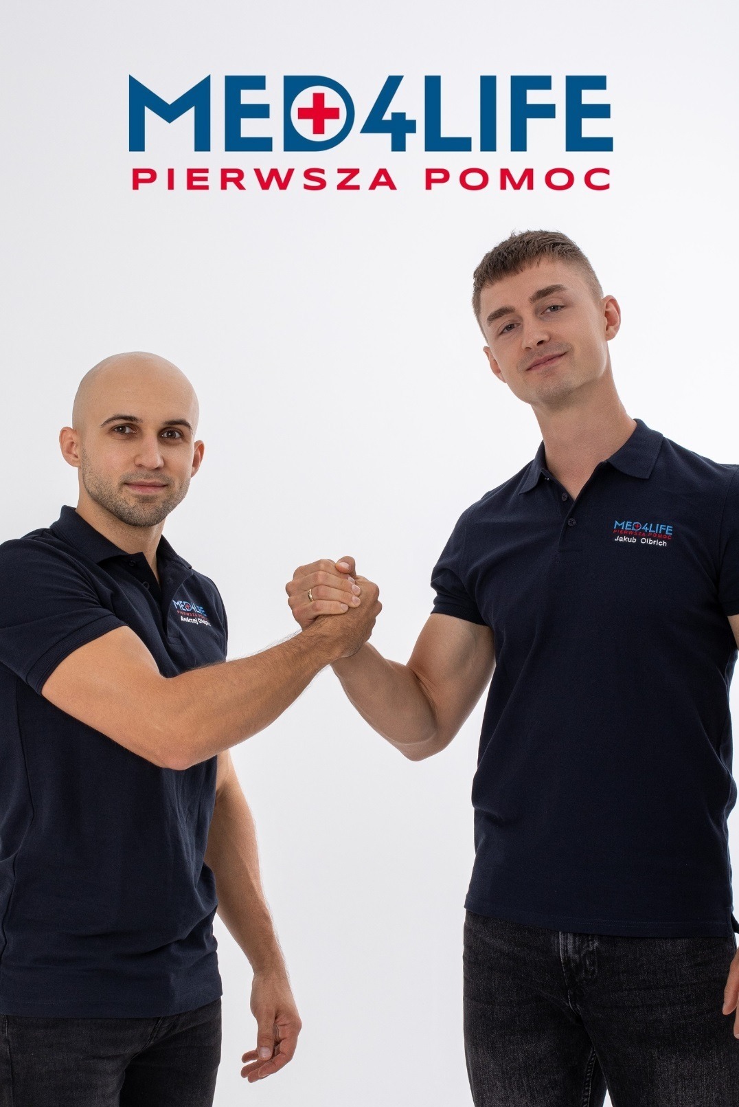 Dwóch mężczyzn w granatowych koszulkach polo z logo Med4Life ściska sobie dłonie na białym tle, widoczny napis PIERWSZA POMOC nad nimi.