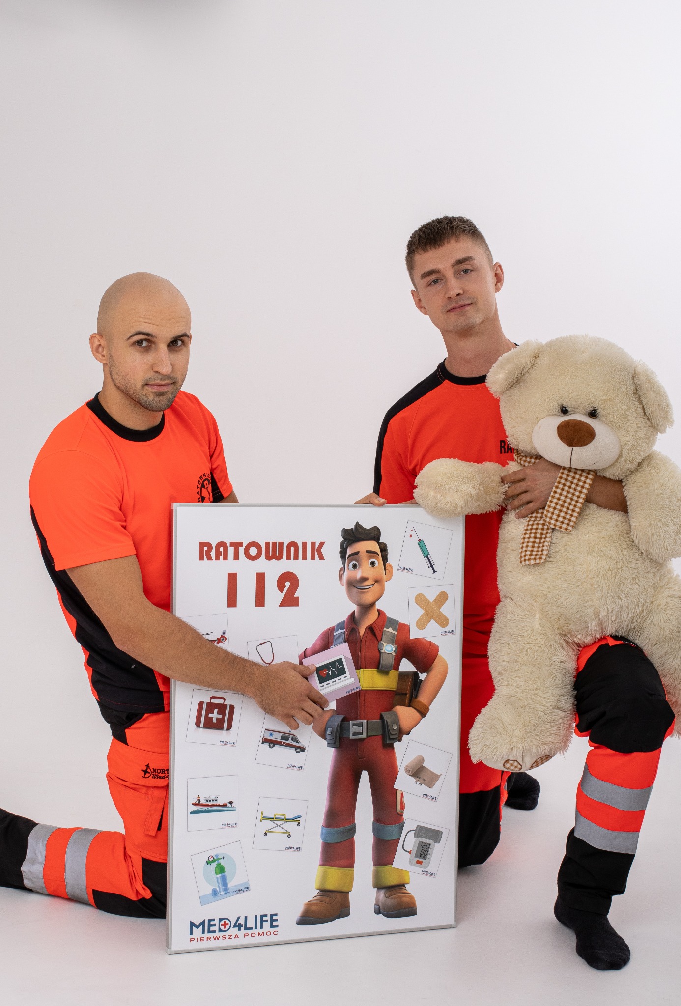 Dwóch ratowników medycznych w pomarańczowych uniformach pozuje z pluszowym misiem i planszą edukacyjną z napisem 'Ratownik 112' oraz ikonami sprzętu medycznego i opatrunków.