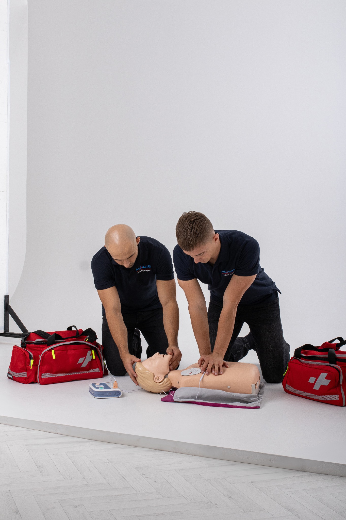 Dwóch mężczyzn w granatowych koszulkach demonstruje resuscytację krążeniowo-oddechową na manekinie, obok czerwone torby medyczne i defibrylator na białym tle.
