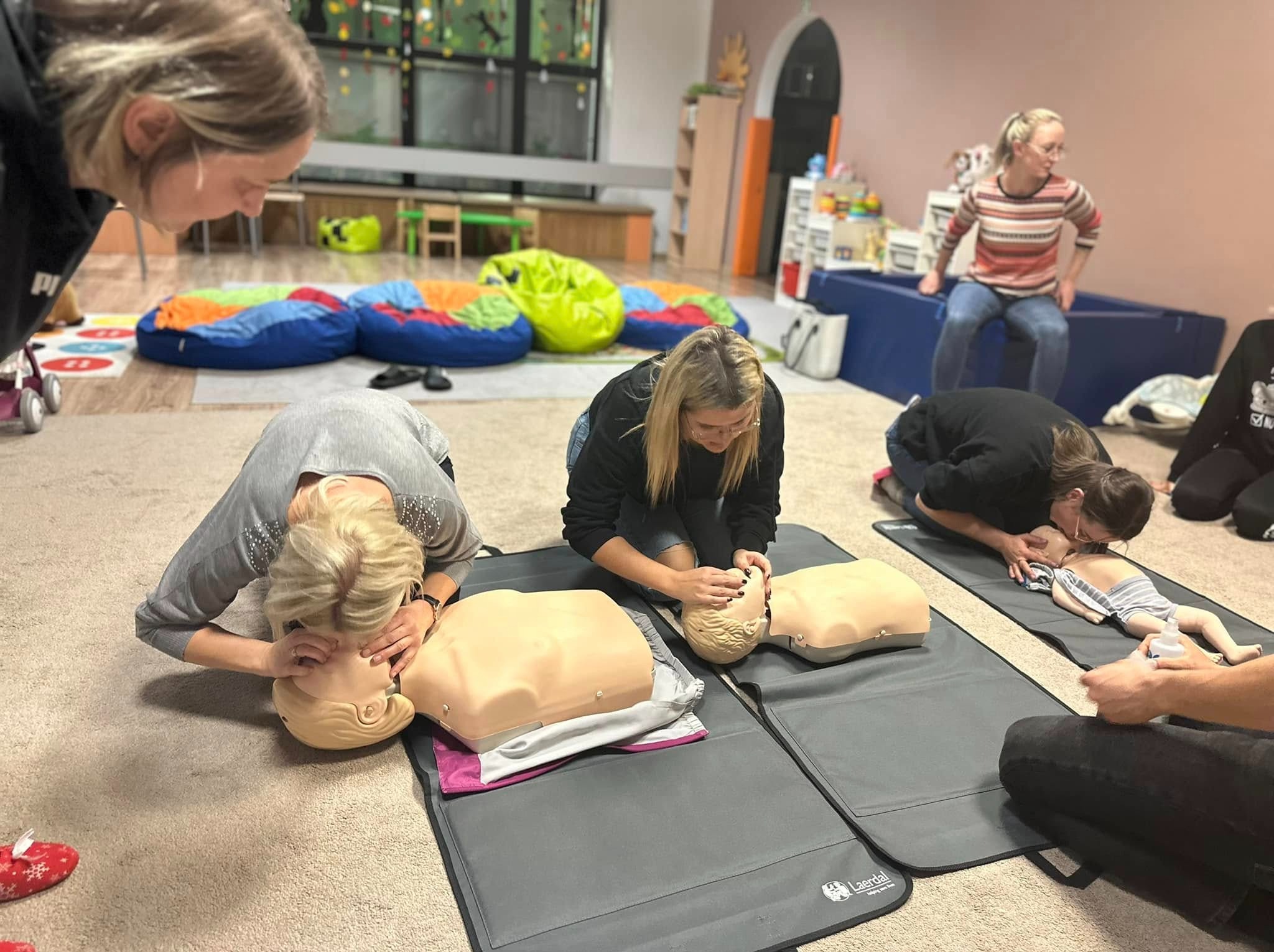 Uczestnicy kursu ćwiczą resuscytację na manekinach niemowląt i dorosłych na podłodze w sali szkoleniowej z kolorowymi pufami w tle.
