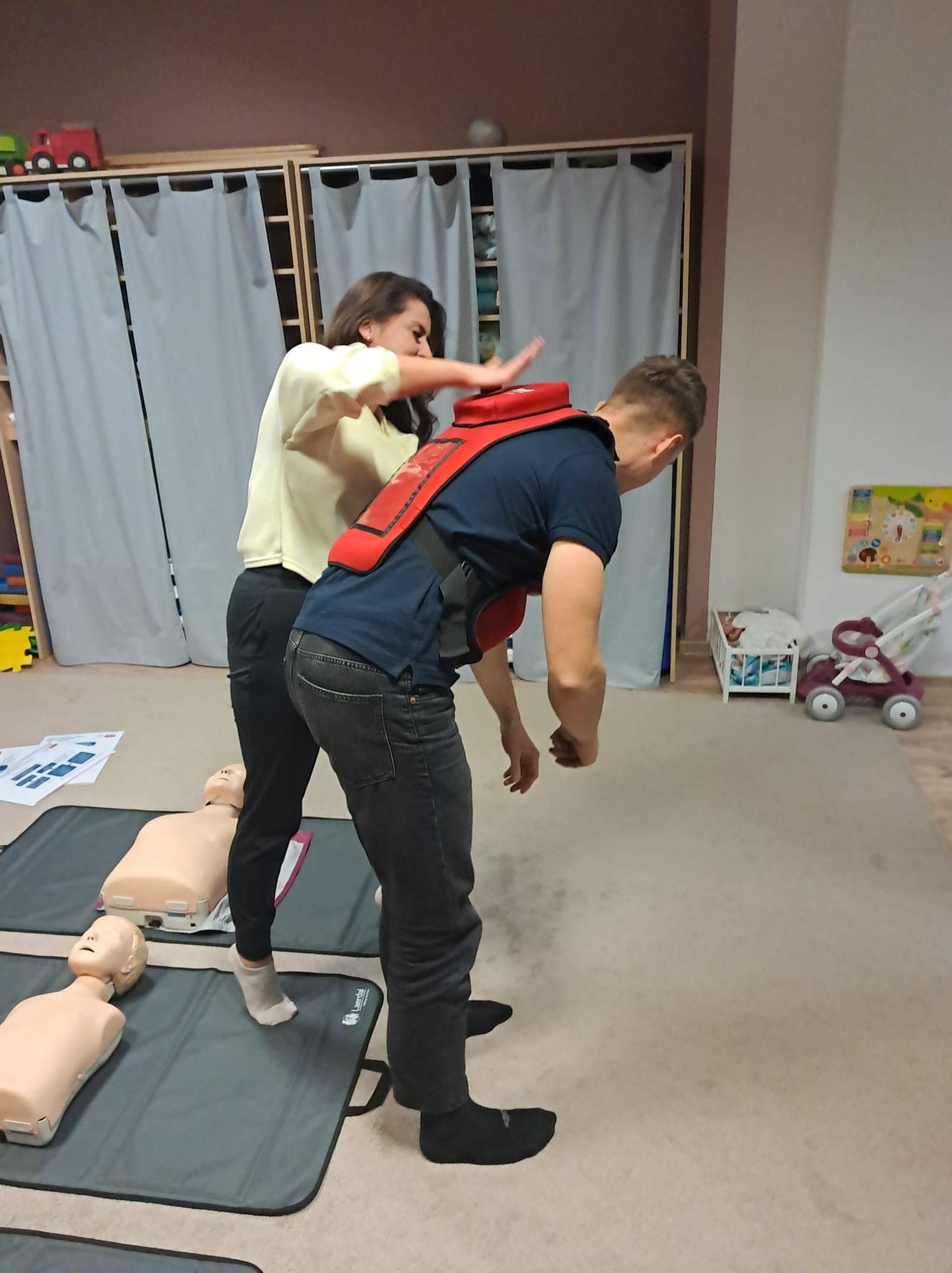 Instruktorka demonstruje uciskanie klatki piersiowej na manekinie z założonym urządzeniem do nauki resuscytacji, w tle widoczne manekiny na matach i dziecięce zabawki.