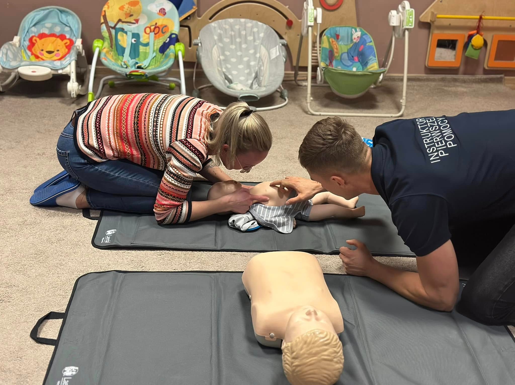 Instruktorzy udzielają pierwszej pomocy niemowlęciu na manekinie podczas szkolenia, w tle widoczne dziecięce huśtawki i zabawki.