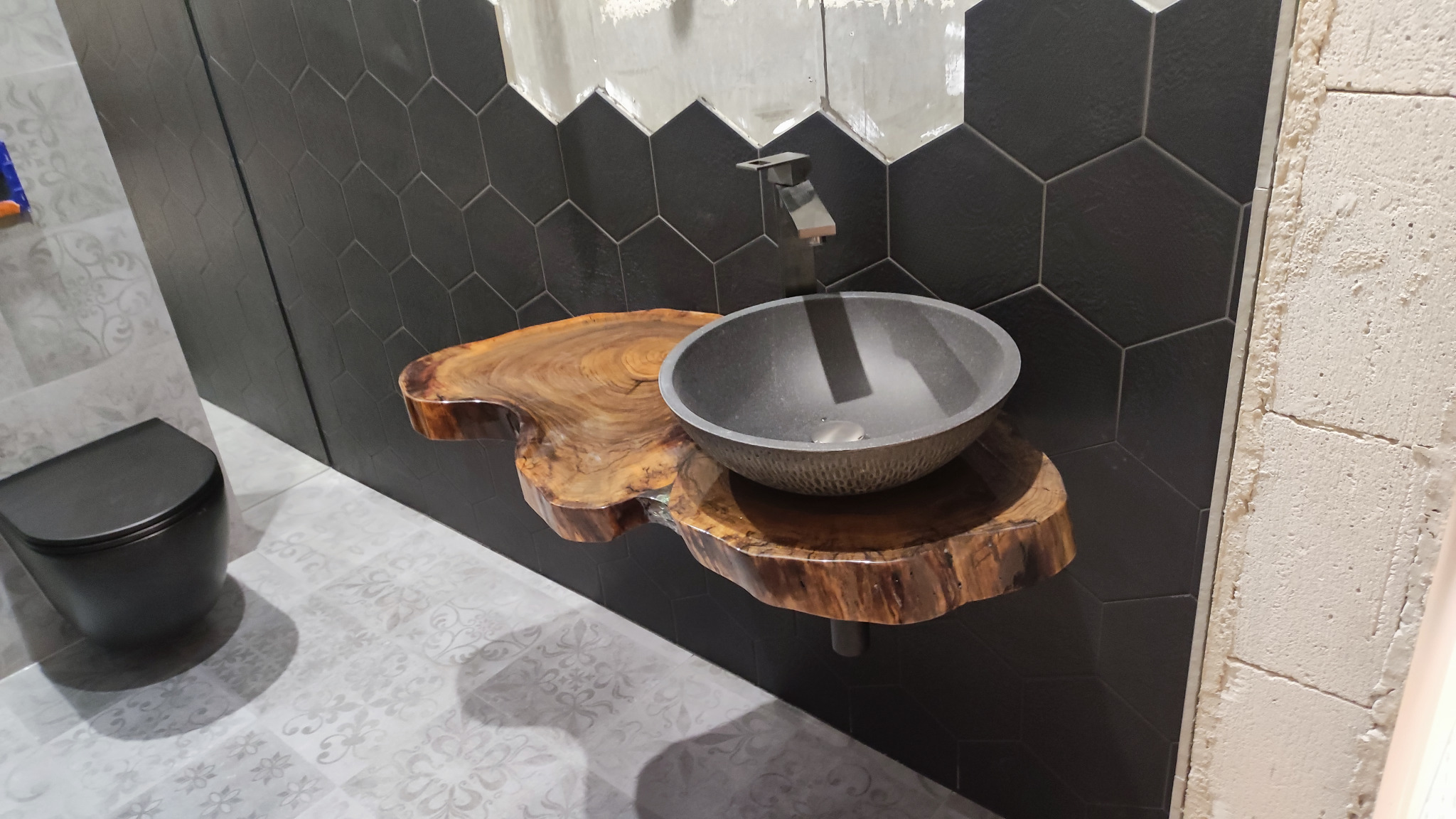 Nowoczesna łazienka z umywalką w kształcie misy, osadzoną na blacie z naturalnego drewna, czarne heksagonalne płytki na ścianie, wisząca toaleta w tle oraz dekoracyjna podłoga.