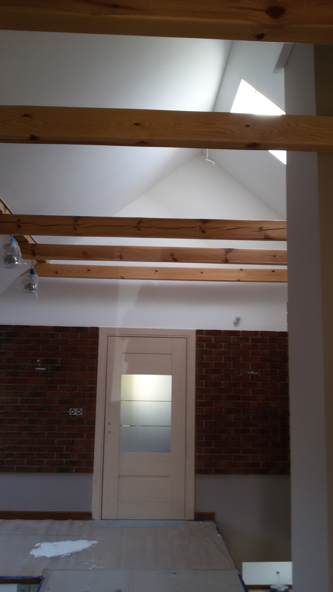 Wnętrze w trakcie remontu: surowa ceglana ściana, białe drzwi z przeszkleniem, drewniane belki stropowe, zabezpieczona podłoga z plamą farby, widoczne okno dachowe.