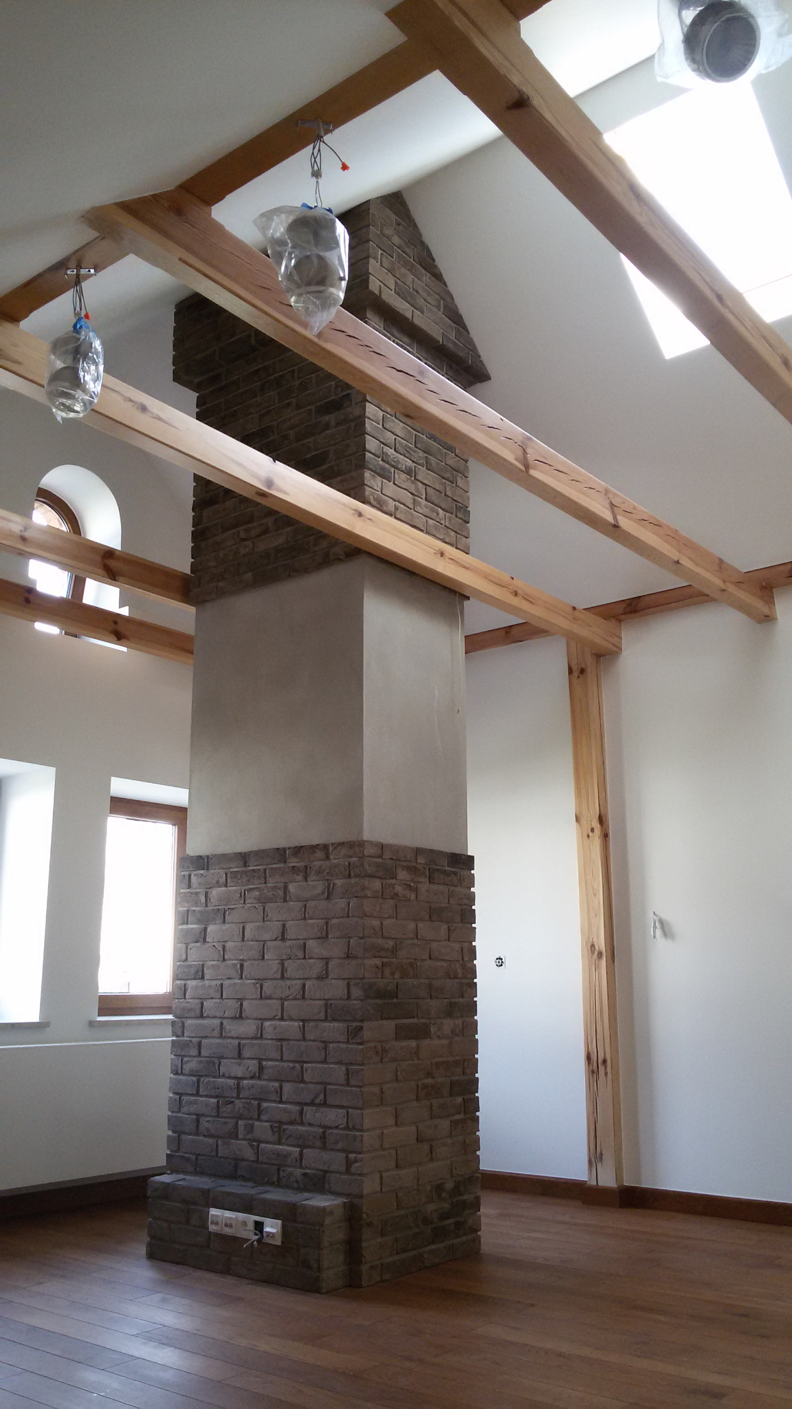 Wnętrze w trakcie wykańczania: murowany komin z cegły przechodzi w gładź, drewniane belki stropowe, podłoga z parkietu, okna z łukami, lampy zabezpieczone folią.