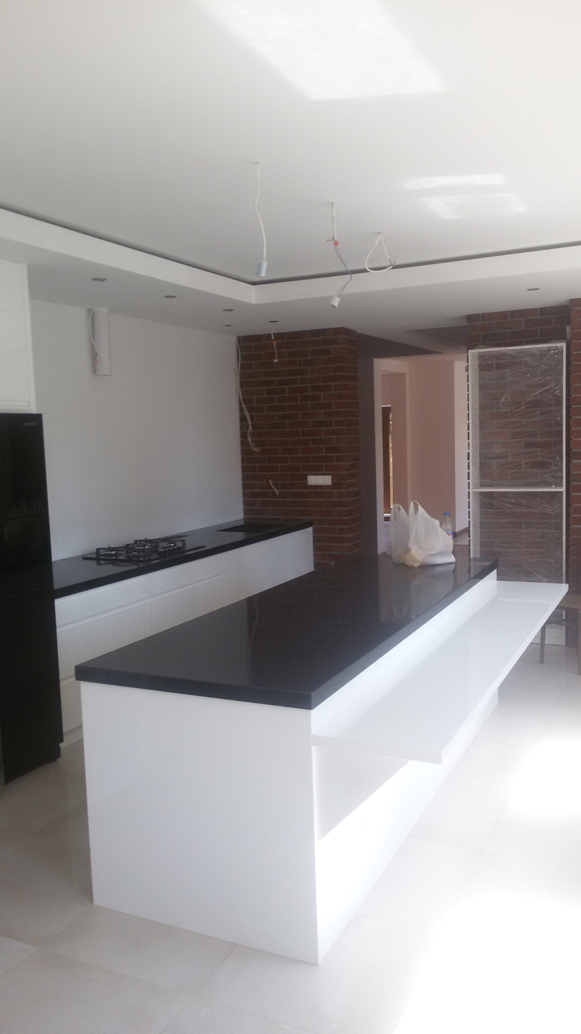 Wnętrze kuchni w trakcie wykańczania z wyspą z czarnym blatem i białym korpusem, widoczne przewody oświetleniowe, ściana z cegły klinkierowej w tle.