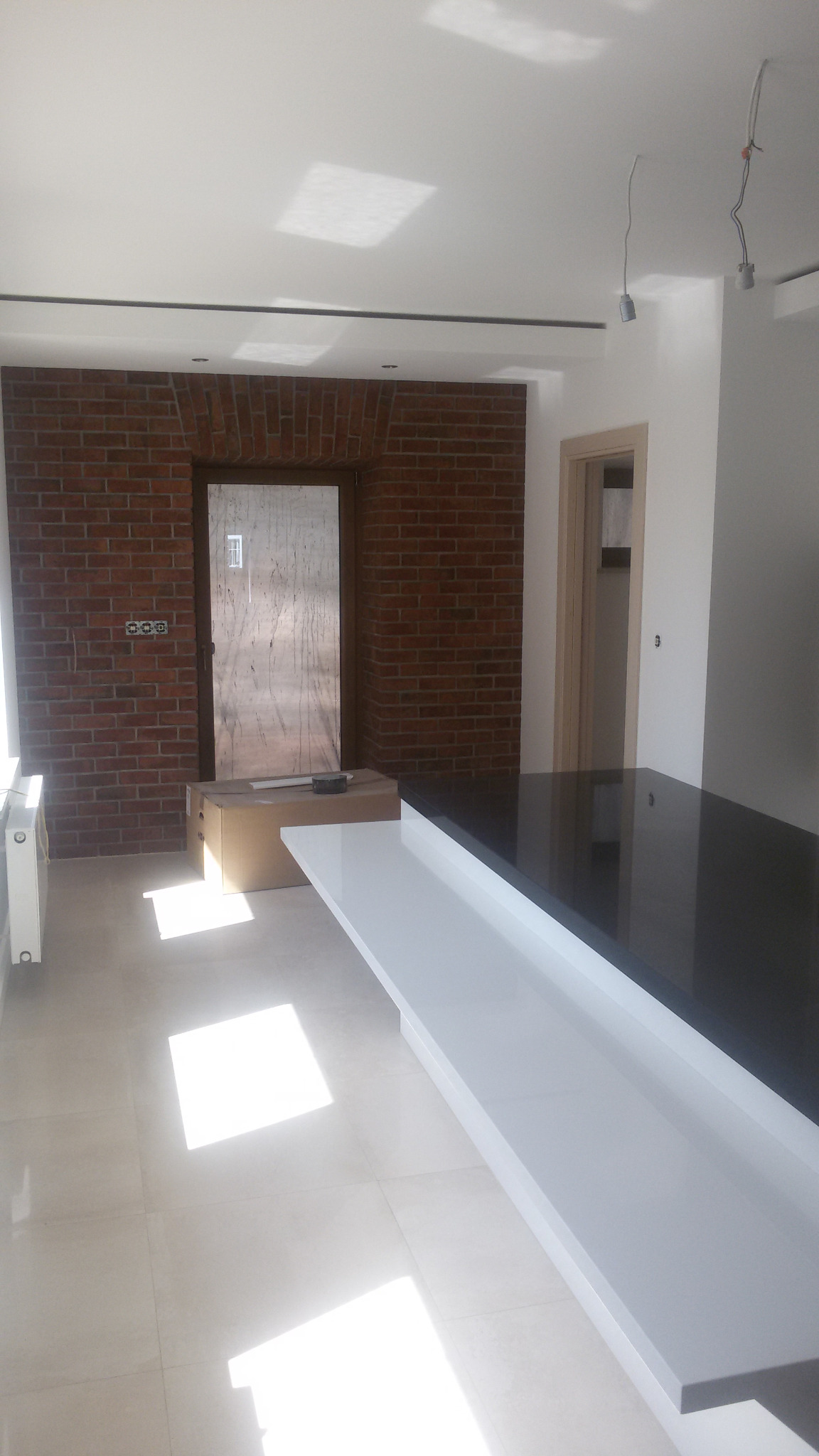 Wnętrze w trakcie prac w Krzeszowicach: ściana z cegły z drzwiami zabezpieczonymi folią, biała wyspa kuchenna z czarnym blatem, widoczne niedokończone podłączenia oświetlenia sufitowego.