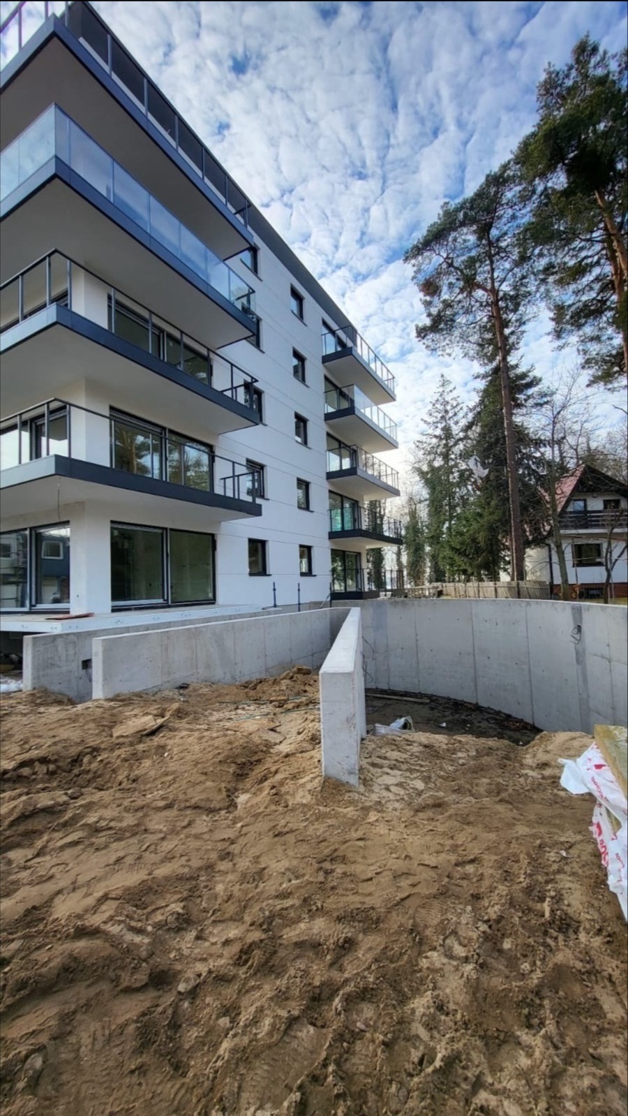 Nowoczesny budynek apartamentowy z balkonami i szklanymi balustradami, widoczny z poziomu terenu z wykopem i betonowym fundamentem w trakcie budowy.