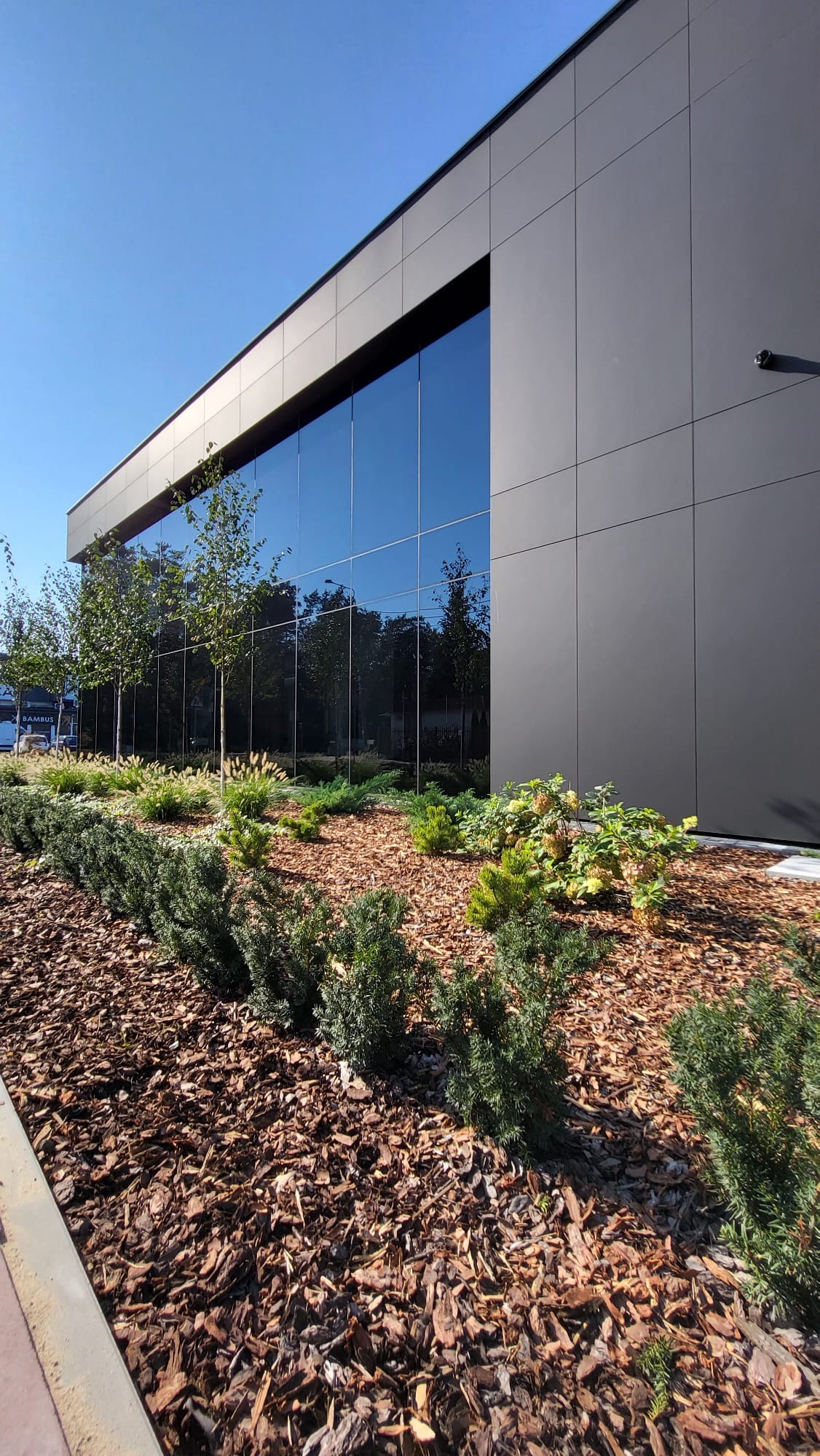 Nowoczesny budynek usługowy z fasadą z ciemnych paneli i dużymi oknami, otoczony zielenią i korą drzewną.