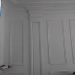 Jacek - Biała ściana z dekoracyjnymi panelami i sztukaterią, widoczny fragment sufitu z ozdobnym elementem, minimalistyczny design.