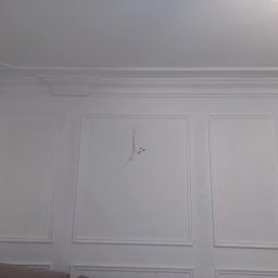 Jacek - Biała ściana z dekoracyjnymi panelami i listwami przysufitowymi, widoczne kable wystające ze ściany, fragment sofy w dolnej części kadru.