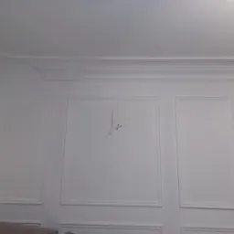 Biała ściana z dekoracyjnymi panelami i listwami przysufitowymi, widoczne kable wystające ze ściany, fragment sofy w dolnej części kadru.