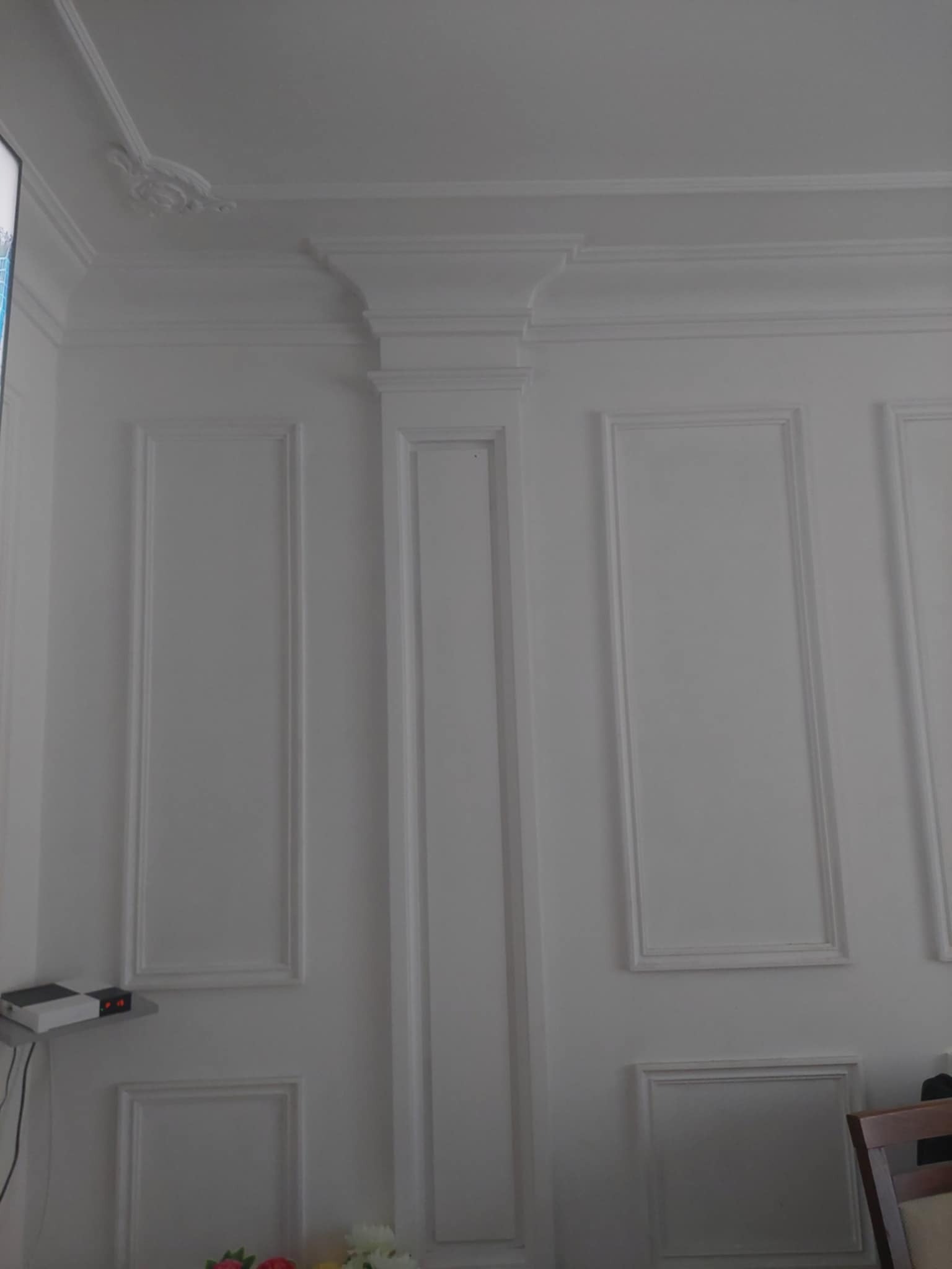 Biała ściana z dekoracyjnymi panelami i sztukaterią, widoczny fragment sufitu z ozdobnym elementem, minimalistyczny design.