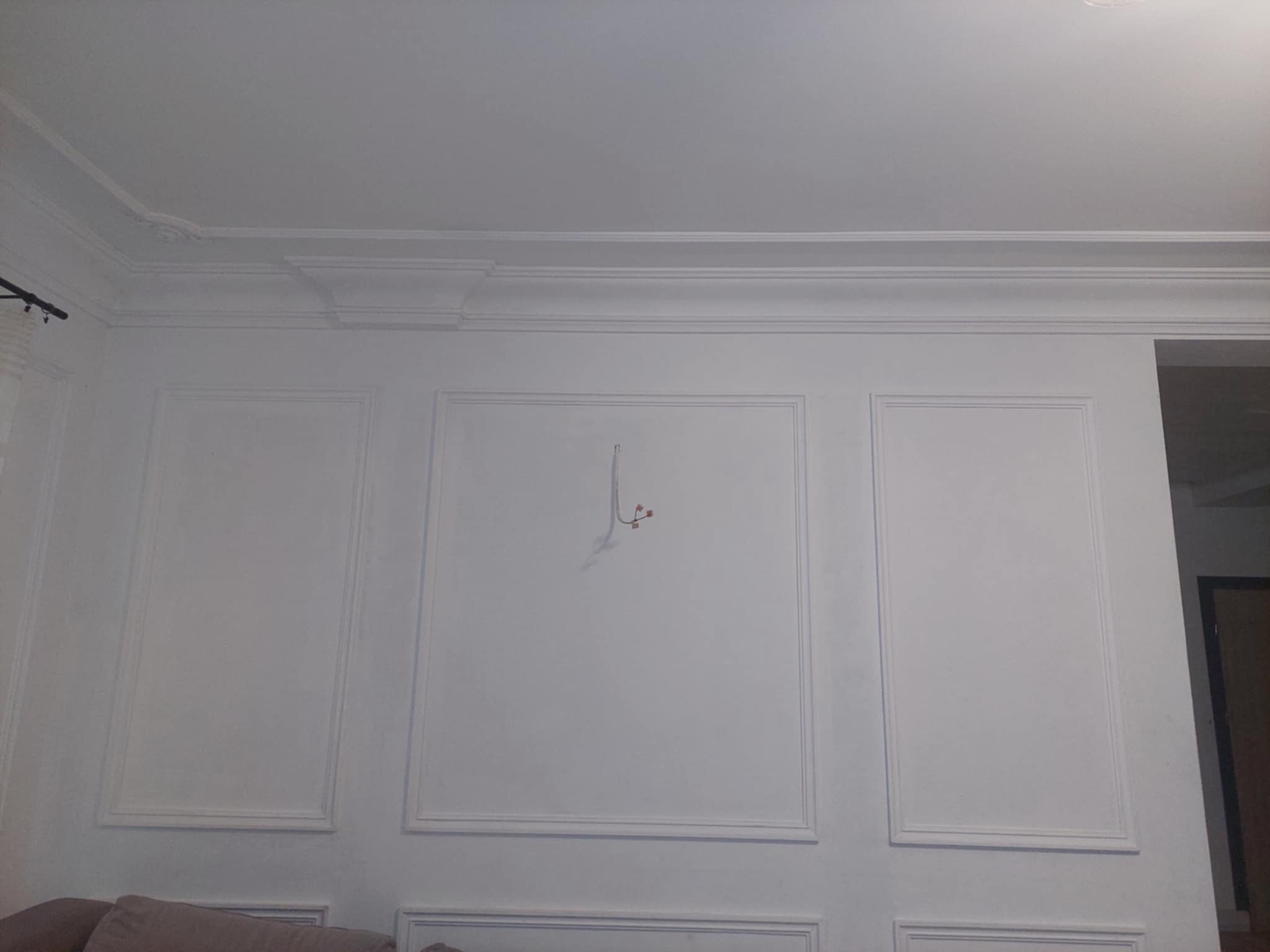 Biała ściana z dekoracyjnymi panelami i listwami przysufitowymi, widoczne kable wystające ze ściany, fragment sofy w dolnej części kadru.