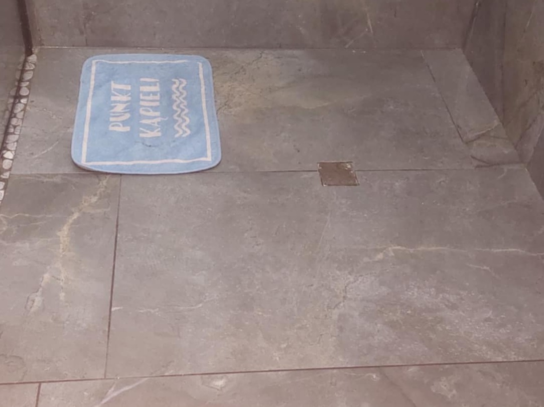 Szare płytki podłogowe w łazience z odpływem liniowym i błękitnym dywanikiem z napisem 'Punkt Kąpieli'.