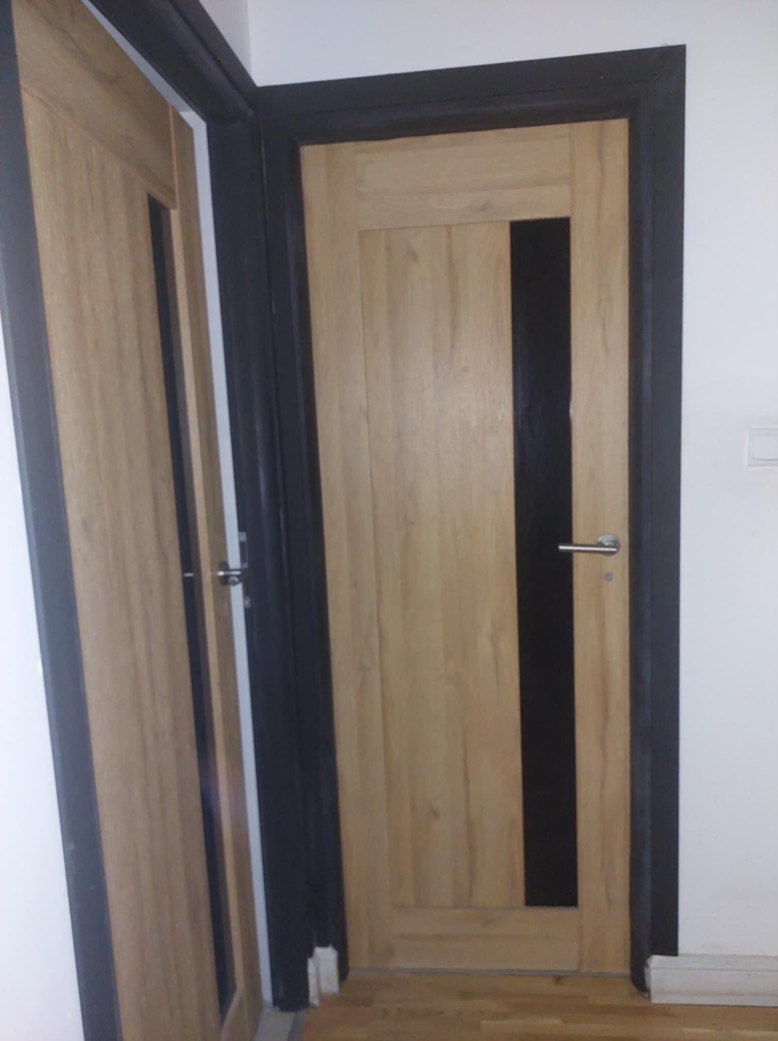 Zamknięte, drewnopodobne drzwi wewnętrzne z czarnym pionowym pasem, osadzone w czarnej futrynie, widoczne z boku, na tle białej ściany z włącznikiem.